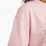 A young woman taking a mirror selfie wearing a VALE GIRL graphic t-shirt made of breathable cotton with a floral design, available in pink, light blue, and maroon.