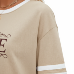 Woman wearing a VALE branded varsity t-shirt made of breathable cotton with white sleeve stripes and a vintage filigree logo, available in beige, olive, and brown.