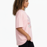 A young woman taking a mirror selfie wearing a VALE GIRL graphic t-shirt made of breathable cotton with a floral design, available in pink, light blue, and maroon.