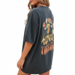 Rear and front view of a woman wearing an oversized breathable cotton t-shirt in charcoal, beige, and brown. The back features a colorful retro graphic of mushrooms and daisies with the text "Love Yourself." The front features a small white minimalist logo on the left chest.