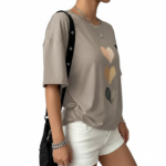 Front view of a woman wearing an oversized breathable cotton t-shirt with a triple heart graphic design and a small "V" logo, styled with white denim shorts and a black shoulder bag.