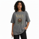 A woman modeling an oversized VALE brand t-shirt featuring a vintage-style Protea flower graphic and celestial symbols. Available in black, grey, and cream colors.