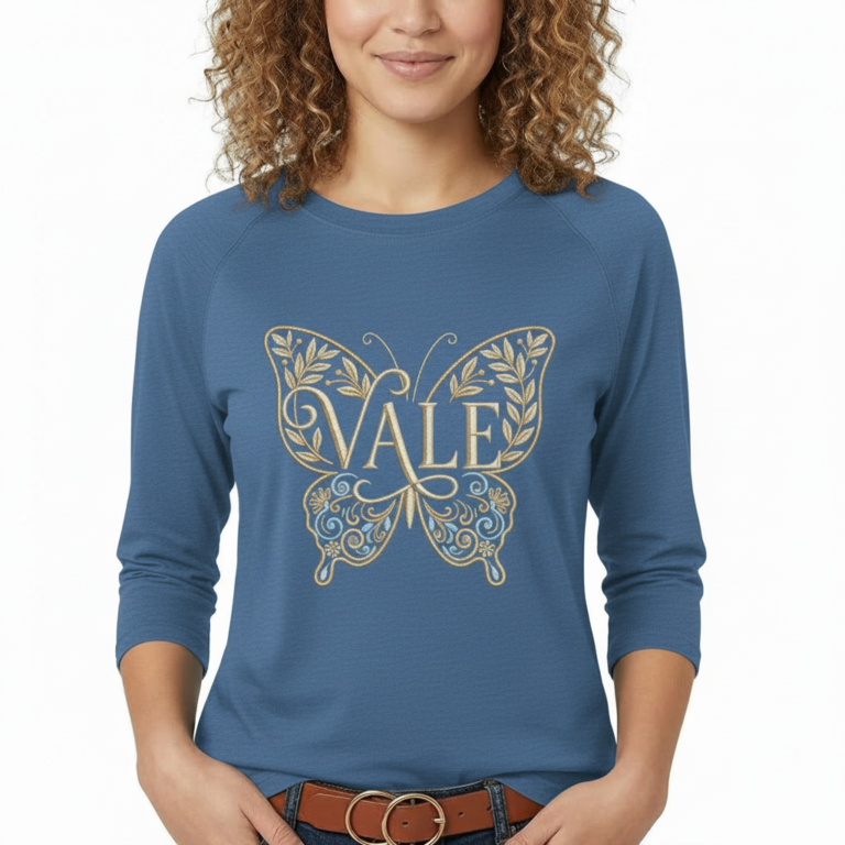 A woman wearing a VALE brand 3/4 sleeve raglan shirt made of breathable cotton, featuring a large, ornate butterfly embroidery on the chest in gold and light blue.