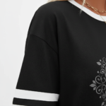 A person wearing a breathable cotton VALE graphic t-shirt with varsity stripes on sleeves, available in black, brown, and orange.