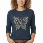 A woman wearing a VALE brand 3/4 sleeve raglan shirt made of breathable cotton, featuring a large, ornate butterfly embroidery on the chest in gold and light blue.