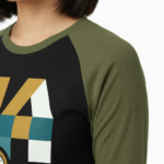 A woman wearing a breathable cotton raglan baseball shirt featuring a colorful geometric "VALE" logo and a central compass graphic. Available in Maroon, Grey, and Olive Green sleeves with a black or burgundy torso.