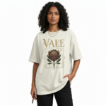 A woman modeling an oversized VALE brand t-shirt featuring a vintage-style Protea flower graphic and celestial symbols. Available in black, grey, and cream colors.
