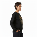 Person wearing a VALE graphic cotton sweatshirt featuring botanical leaves, acorns, and stars in Black, Forest Green, and Burnt Orange colors.