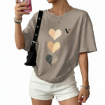 Front view of a woman wearing an oversized breathable cotton t-shirt with a triple heart graphic design and a small "V" logo, styled with white denim shorts and a black shoulder bag.