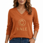 Front view of a woman wearing a VALE brand long sleeve V-neck cotton shirt featuring a gold celestial sun and moon logo, available in maroon, brown, and orange.