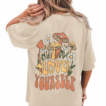 Rear and front view of a woman wearing an oversized breathable cotton t-shirt in charcoal, beige, and brown. The back features a colorful retro graphic of mushrooms and daisies with the text "Love Yourself." The front features a small white minimalist logo on the left chest.