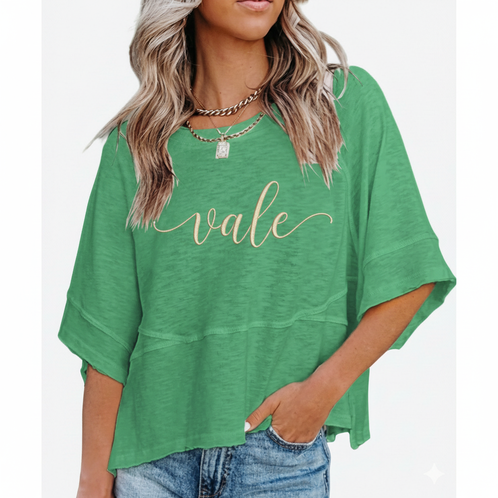 Woman wearing a breathable cotton oversized "Vale" t-shirt with gold script lettering and horizontal seam detail, available in green, blue, and red.