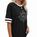 A person wearing a breathable cotton VALE graphic t-shirt with varsity stripes on sleeves, available in black, brown, and orange.