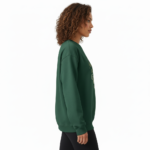 Woman wearing a VALE graphic cotton sweatshirt featuring a swallow, butterfly, and botanical nature design in Forest Green, Black, and Purple colors.