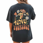 Rear and front view of a woman wearing an oversized breathable cotton t-shirt in charcoal, beige, and brown. The back features a colorful retro graphic of mushrooms and daisies with the text "Love Yourself." The front features a small white minimalist logo on the left chest.