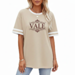 Woman wearing a VALE branded varsity t-shirt made of breathable cotton with white sleeve stripes and a vintage filigree logo, available in beige, olive, and brown.