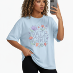A young woman taking a mirror selfie wearing a VALE GIRL graphic t-shirt made of breathable cotton with a floral design, available in pink, light blue, and maroon.