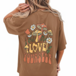 Rear and front view of a woman wearing an oversized breathable cotton t-shirt in charcoal, beige, and brown. The back features a colorful retro graphic of mushrooms and daisies with the text "Love Yourself." The front features a small white minimalist logo on the left chest.