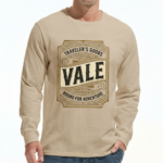 A man wearing a VALE Traveler’s Goods long-sleeve cotton shirt featuring a vintage-style gold and cream graphic label that says "Bound for Adventure" on the chest.