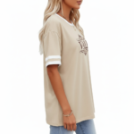 Woman wearing a VALE branded varsity t-shirt made of breathable cotton with white sleeve stripes and a vintage filigree logo, available in beige, olive, and brown.
