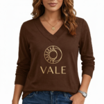 Front view of a woman wearing a VALE brand long sleeve V-neck cotton shirt featuring a gold celestial sun and moon logo, available in maroon, brown, and orange.
