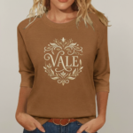 Woman wearing a breathable cotton 3/4 sleeve raglan shirt featuring a gold-toned floral "VALE" crest design, available in Navy Blue, Earthy Brown, and Maroon.