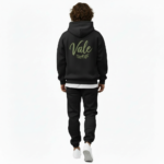 A man wearing a premium breathable cotton tracksuit featuring a sage green "Vale" script logo and olive branch graphic on the back, available in Black, Charcoal Grey, and Burgundy.