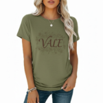 A woman wearing a slim-fit cotton graphic t-shirt featuring the word "VALE" inside a floral wreath design. Available in Dusty Blue, Olive Green, and Deep Maroon colors.