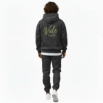 A man wearing a premium breathable cotton tracksuit featuring a sage green "Vale" script logo and olive branch graphic on the back, available in Black, Charcoal Grey, and Burgundy.