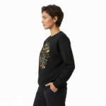 Person wearing a VALE graphic cotton sweatshirt featuring botanical leaves, acorns, and stars in Black, Forest Green, and Burnt Orange colors.