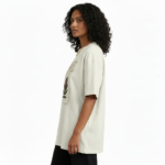 A woman modeling an oversized VALE brand t-shirt featuring a vintage-style Protea flower graphic and celestial symbols. Available in black, grey, and cream colors.