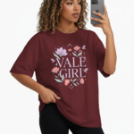 A young woman taking a mirror selfie wearing a VALE GIRL graphic t-shirt made of breathable cotton with a floral design, available in pink, light blue, and maroon.