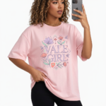 A young woman taking a mirror selfie wearing a VALE GIRL graphic t-shirt made of breathable cotton with a floral design, available in pink, light blue, and maroon.