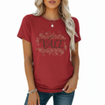 A woman wearing a slim-fit cotton graphic t-shirt featuring the word "VALE" inside a floral wreath design. Available in Dusty Blue, Olive Green, and Deep Maroon colors.